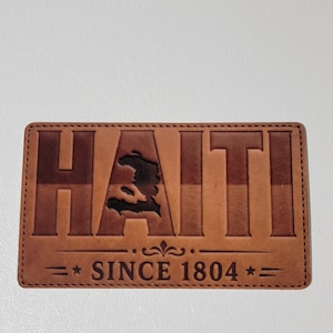 May include: A rectangular, brown leather patch with the word "HAITI" embossed in large, capital letters. The country's outline is cut out within the letters. Below, it reads "SINCE 1804" with star accents.