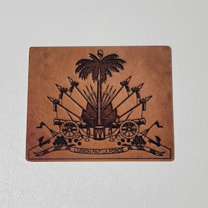 May include: A rectangular, brown leather item featuring a detailed emblem. The emblem includes a palm tree, flags, cannons, and the text "L'UNION FAIT LA FORCE". The design is intricately etched into the leather.