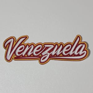 May include: A white, red, and yellow Venezuela sticker. The word "Venezuela" is written in a cursive font with a white fill, red outline, and yellow shadow. The sticker is rectangular and has a slight 3D effect.