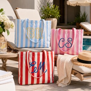 May include: Three canvas tote bags with vertical stripes in light blue, pink, and red. Each bag features two shoulder straps and a large embroidered monogram on the front. The blue bag displays yellow letters BR, the pink bag has blue letters CS, and the red bag shows blue letters EM.