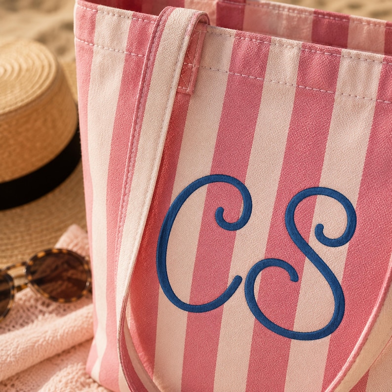 May include: A tote bag featuring vertical stripes in alternating shades of light pink and dark pink. The front of the bag is embroidered with the large, dark blue cursive letters CS in a stylized monogram font, with two matching fabric shoulder straps.