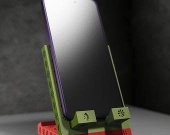 May include: A red, green, and purple phone stand with a phone in place. The stand has a Christmas theme with snowflake and tree cutouts. The phone has a black screen and a purple border.