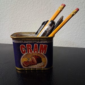 May include: A rectangular metal tin with a blue exterior and the word CRAM printed in red letters above an illustration of sliced meat. The tin serves as a holder for two yellow wooden pencils with pink erasers and two black ink pens.