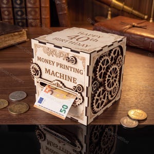 Könnte beinhalten: Eine helle Holzbox mit lasergeschnittenen Zahnrädern an der Seite und dem Text "HAPPY 40th BIRTHDAY" sowie "MONEY PRINTING MACHINE", der oben und vorne eingraviert ist. Ein Fünfzig-Euro-Schein steckt teilweise in einem Schlitz an der Vorderseite.