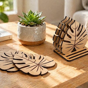 May include: A set of light-colored wooden coasters shaped like monstera leaves, featuring dark laser-cut vein details. The set includes a matching wooden holder with a slatted base, and two individual coasters are placed flat on the surface.
