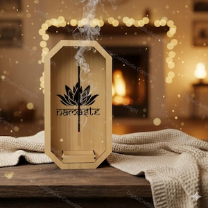 以下が含まれることがあります： 蓮の花のデザインと「namaste」の文字が黒で描かれた木製のお香立て。お香から煙が立ち上り、穏やかな雰囲気を作り出しています。お香立ては木製のテーブルの上にあり、背景には柔らかな毛布があります。