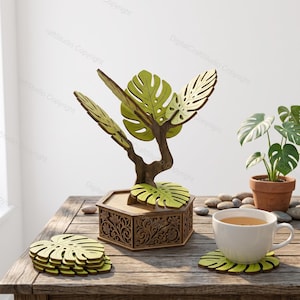 Könnte beinhalten: Ein hölzerner Untersetzerhalter in Baumform mit hellgrünen, monstera-blattförmigen Untersetzern. Der sechseckige Sockel ist mit floralen Schnitzereien verziert. Ein Stapel passender Blattuntersetzer liegt links, einer steht unter einer weißen Tasse.