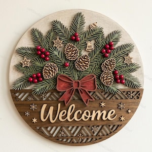 May include: A circular wooden wall sign featuring a layered design with a light-coloured top half and a dark-coloured bottom half. The sign displays the word "Welcome" in script, decorated with green pine branches, red berries, pinecones, and stars.