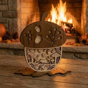 Op de afbeelding: Een gelaagde houten decoratie in de vorm van een eikel met een donkerbruine bovenkant met uitgesneden eikels en takken, en een lichtbeige onderkant met gedetailleerde lasergesneden ontwerpen van een muis, diverse paddenstoelen, bladeren en eikels op een bladvormige voet.