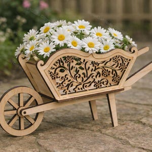 Può includere: Una fioriera a carriola in legno piena di margherite bianche con centri gialli. La carriola ha un design decorativo floreale intagliato sulla parte anteriore e sui lati. La fioriera è in legno chiaro e si trova su una superficie di pietra.