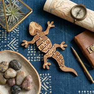Puede incluir: Una figura de lagarto de madera con intrincados patrones grabados con láser en el lomo y partes del cuerpo segmentadas. El lagarto está colocado sobre una tela azul estampada junto a una brújula de latón, una planta de aire y piedras.