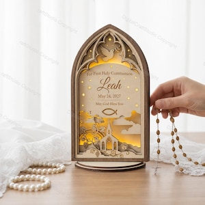 Puede incluir: Un recuerdo de Primera Comunión de madera en forma de arco con el nombre "Leah" y la fecha 24 de mayo de 2027. El diseño incluye una paloma, una iglesia y un símbolo de pez, con el texto "May God Bless You". Un rosario se sostiene cerca.