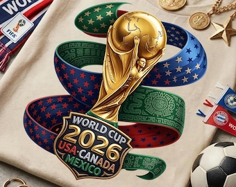 Op de afbeelding: Een beige t-shirt met een gouden trofee met een wereldbol en de tekst "World Cup 2026 USA-Canada-Mexico". Een voetbal, een sjaal en gouden sieraden zijn ook zichtbaar. Linten met sterren en Azteekse patronen wikkelen zich om de trofee.