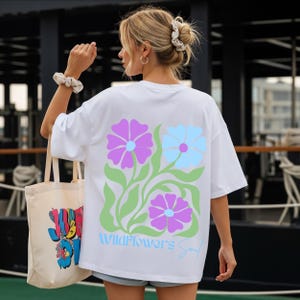 Könnte beinhalten: Weißes T-Shirt mit einem floralen Design in Lila und Hellblau, begleitet von grünen Blättern. Der Text "Wildflowers Soul" ist unter den Blumen in hellblauer Schrift gedruckt. Eine beige Tragetasche mit bunten Grafiken ist ebenfalls zu sehen.