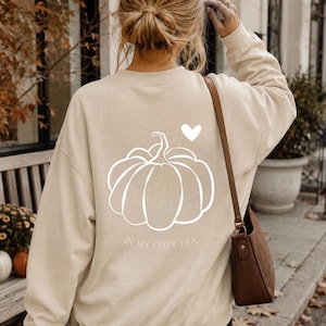 Könnte beinhalten: Ein beigefarbenes Langarm-Sweatshirt mit Rundhalsausschnitt und einer weißen Linienzeichnung eines Kürbisses auf der Rückseite. Über dem Kürbis befindet sich ein kleines weißes Herz und darunter der weiße Schriftzug IN MY COZY ERA.