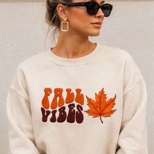 Könnte beinhalten: Ein cremefarbenes Sweatshirt mit Rundhalsausschnitt, das den Schriftzug FALL VIBES in einer retro-gewellten Schriftart mit orangefarbenen und dunkelbraunen Buchstaben sowie ein gesticktes orangefarbenes Ahornblatt auf der Vorderseite zeigt.