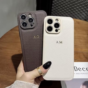 Peut inclure: Deux coques de téléphone en cuir, une marron et une blanc cassé, chacune avec un monogramme doré. La coque marron porte "A.O" et la coque blanc cassé porte "A.M". Les coques ont une finition texturée.