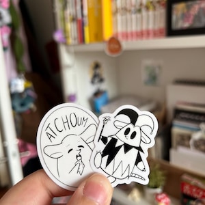 Puede incluir: Dos pegatinas de vinilo troqueladas con ilustraciones de líneas en blanco y negro. Una pegatina circular muestra a un personaje estornudando con el texto "ATCHOUM" encima. La otra muestra a un personaje con un gorro de bufón.