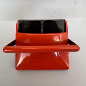 Puede incluir: Un organizador de escritorio de color naranja brillante con acabado satinado. El artículo presenta una base rectangular con un asa horizontal ancha que se extiende desde el frente. El interior está dividido en dos compartimentos con forro negro.