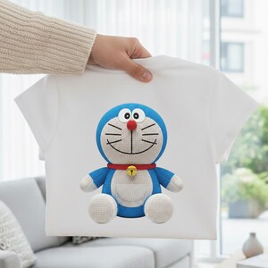May include: White t-shirt featuring a blue cartoon character with a red nose, bell, and collar. The character has a blue head, arms, and body with white accents. The t-shirt is held up by a person.