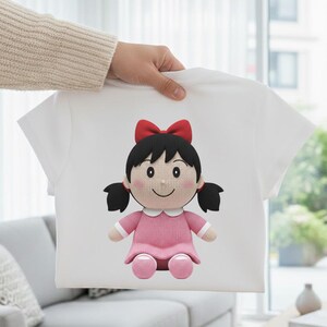 May include: White t-shirt with a cartoon doll design. The doll has black hair, a red bow, and a pink dress. The doll's face is smiling. The shirt is held by a person.