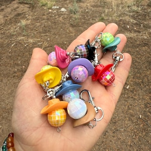 May include: A collection of colorful cowboy hat keychains with disco ball accents. The keychains feature a variety of colors, including yellow, pink, blue, and red. Each keychain has a silver clasp.