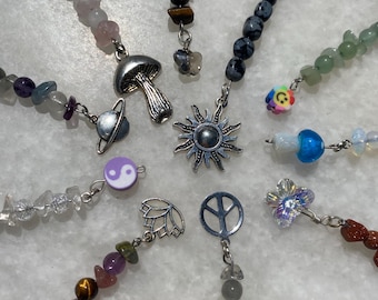 Puede incluir: Una colección de dijes de cuentas con diversas piedras naturales y colgantes de metal que incluyen un hongo, un sol, un símbolo de paz, una flor de loto, un símbolo yin-yang, un planeta, una flor y una cara sonriente, unidos a un anillo.