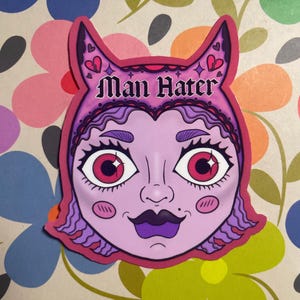 May include: A sticker featuring a stylized illustration of a woman's face with the text "Man Hater". The face is pink with purple hair, a dark purple mustache, and large eyes with red irises. The sticker has a pink border and is set against a floral background.