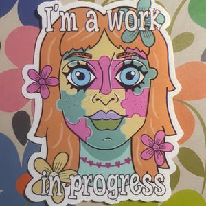 Puede incluir: Una pegatina con una colorida ilustración del rostro de una mujer compuesta por piezas de rompecabezas. La mujer tiene el pelo naranja, ojos azules y flores rosas. El texto "I'm a work in progress" está escrito en la parte superior e inferior de la imagen.