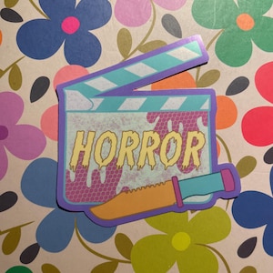 May include: A sticker featuring a film clapperboard design with the word "HORROR" in dripping yellow letters. A knife graphic is included. The sticker has a purple border and is set against a floral background.