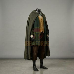 K&ouml;nnte beinhalten: Ein bodenlanger olivgr&uuml;ner Wollumhang mit einer silbernen blattf&ouml;rmigen Schlie&szlig;e, getragen &uuml;ber einer dunkelgr&uuml;nen Jacke, einer senfgelben Weste mit Kn&ouml;pfen und einer braunen Hose. Das Set ist auf einer Schaufensterpuppe vor grauem Hintergrund.