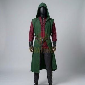 K&ouml;nnte beinhalten: Eine &auml;rmellose gr&uuml;ne Tunika mit Kapuze &uuml;ber einem lang&auml;rmeligen kastanienbraunen Hemd und einer dunklen Hose. Das Outfit umfasst einen braunen Lederg&uuml;rtel mit Metallschnalle, schwarze Handschuhe und eine dunkle Kapuze an einer Schaufensterpuppe.
