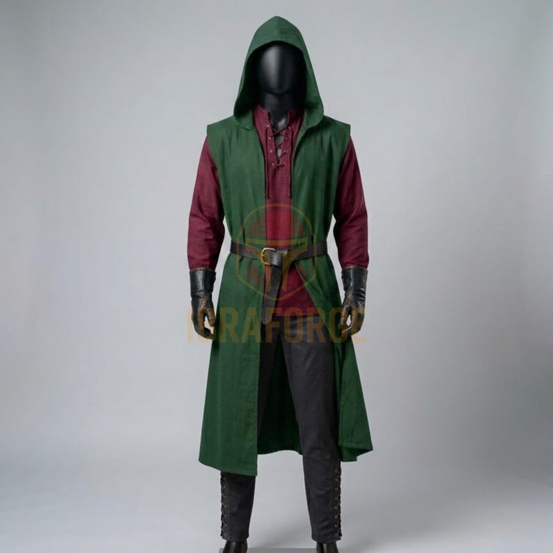 K&ouml;nnte beinhalten: Eine &auml;rmellose gr&uuml;ne Tunika mit Kapuze &uuml;ber einem lang&auml;rmeligen kastanienbraunen Hemd und einer dunklen Hose. Das Outfit umfasst einen braunen Lederg&uuml;rtel mit Metallschnalle, schwarze Handschuhe und eine dunkle Kapuze an einer Schaufensterpuppe.