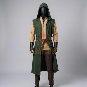 K&ouml;nnte beinhalten: Eine &auml;rmellose, dunkelgr&uuml;ne Tunika mit Kapuze &uuml;ber einem beigen Langarmhemd und einer dunkelbraunen Hose. Das Outfit umfasst einen braunen Lederg&uuml;rtel mit Schnalle in der Taille sowie dunkelbraune Lederhandschuhe an einer Schaufensterpuppe.