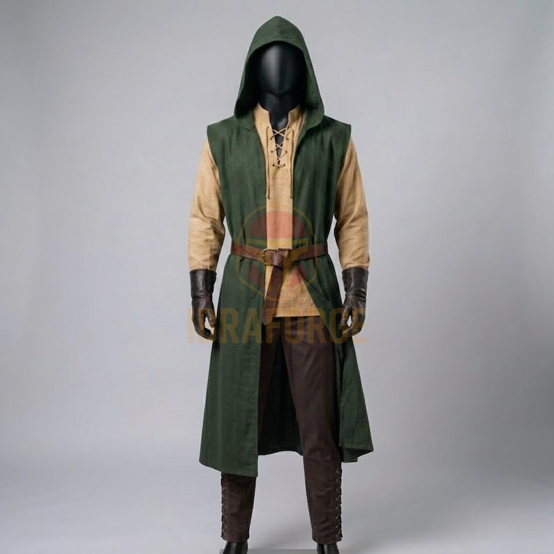 K&ouml;nnte beinhalten: Eine &auml;rmellose, dunkelgr&uuml;ne Tunika mit Kapuze &uuml;ber einem beigen Langarmhemd und einer dunkelbraunen Hose. Das Outfit umfasst einen braunen Lederg&uuml;rtel mit Schnalle in der Taille sowie dunkelbraune Lederhandschuhe an einer Schaufensterpuppe.