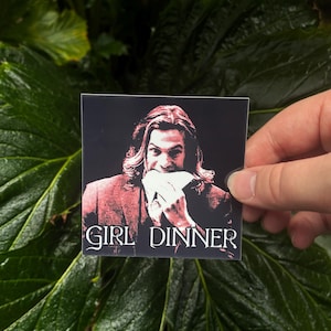 May include: Square sticker with a black background. It features a red-toned image of a person eating, with the white text "GIRL DINNER" below. The sticker is held against a backdrop of large green leaves.