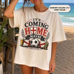 May include: An ivory t-shirt featuring a graphic with the text IT'S COMING HOME...MAYBE, a shield with a red cross, a soccer ball, a glass of beer, a lion, and the text WORLD CUP 2026. The graphic also includes the text COMFORT COLORS 1717 IVORY COLOR.