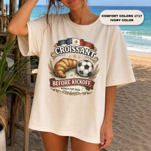 May include: An ivory Comfort Colors 1717 t-shirt featuring a graphic with a croissant, a soccer ball, and a French flag ribbon. The text reads "CROISSANT BEFORE KICKOFF" and "WORLD CUP 2026" in a decorative banner style with wheat accents.