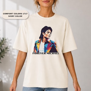 May include: An ivory-colored t-shirt featuring a geometric, multi-colored graphic illustration of Michael Jackson. The design includes the text "FOREVER MICHAEL" in black lettering below the portrait. The shirt has short sleeves and a crew neck.