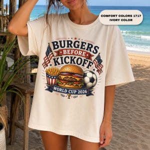 May include: An ivory t-shirt featuring a graphic print with the text "BURGERS BEFORE KICKOFF" and "WORLD CUP 2026". The design includes a hamburger, a container of french fries, a soccer ball, and two American flags. The shirt is Comfort Colors 1717.