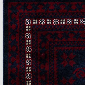 May include: Close-up of a section of a rug with a dark blue and burgundy color scheme. The rug features a geometric pattern with a border of repeating floral motifs. The edge has a fringe of white, red, and blue threads.