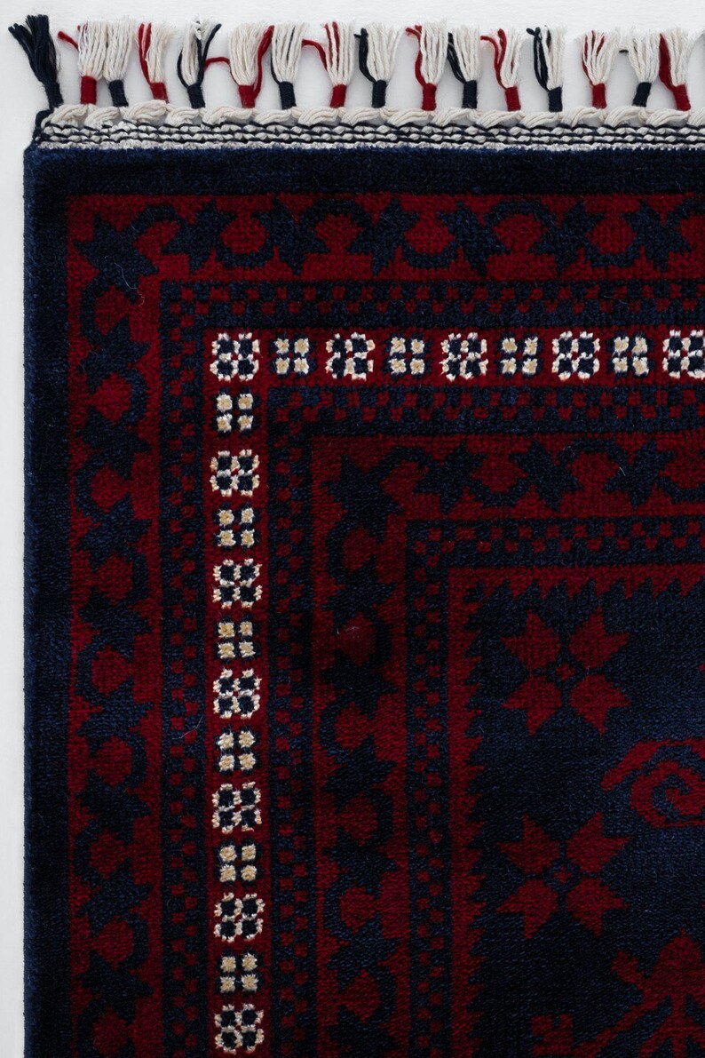 May include: Close-up of a section of a rug with a dark blue and burgundy color scheme. The rug features a geometric pattern with a border of repeating floral motifs. The edge has a fringe of white, red, and blue threads.
