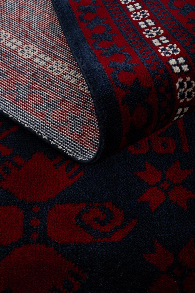 May include: A rolled-up area rug with a dark blue background and a repeating pattern in red and white. The rug's design includes floral and geometric motifs, creating a traditional aesthetic. The edges are finished with a dark blue border.