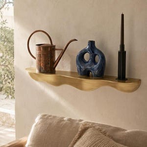 Puede incluir: Una estantería decorativa de latón con borde ondulado sostiene una regadera de cobre, un jarrón de cerámica azul y una vela negra con un soporte negro. La estantería está montada en una pared de color neutro.