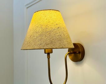 May include: A wall-mounted lamp with a beige fabric shade and a brass-colored metal frame. The lamp has a curved arm and a circular base, mounted on a white wall.