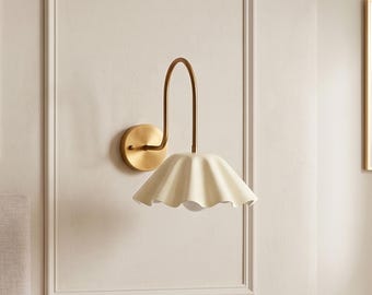 May include: A wall-mounted light fixture featuring a circular brass base and a curved brass arm that supports a cream-colored, scalloped, bell-shaped shade. The design includes a downward-facing light source housed within the fluted metal shade.