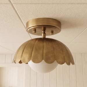 May include: A flush mount ceiling light fitting featuring a brushed brass finish with a scalloped edge dome shade and a central white glass globe. The fitting is attached to a circular brass canopy with two small visible mounting screws.