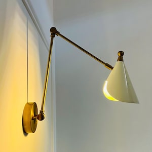 Op de afbeelding: Een aan de muur gemonteerde messing lamp met een witte, conische kap. De lamp heeft een verstelbare arm en een ronde basis. De kap is verlicht en geeft een warm licht af. Het ontwerp is modern en minimalistisch.