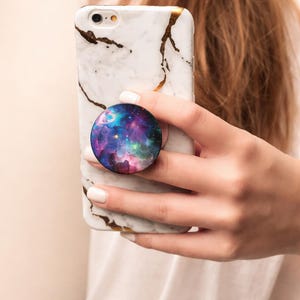 May include: A white marble phone case with gold accents holds a round pop socket. The pop socket features a colorful galaxy design with shades of blue, purple, and pink. The phone is held in a person's hand.