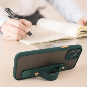 May include: A dark green phone case with a built-in finger strap. The case has a clear back and a dark green border. The strap is attached with a gold-colored button. The phone case is on a desk with a person writing in a notebook.